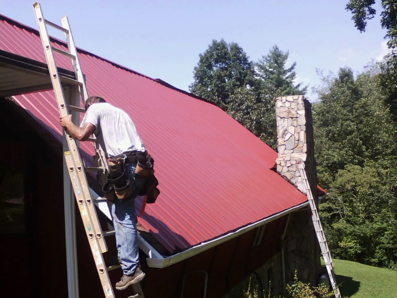 Roofer on a ladder beside a red metal roof and stone chimney — Soffit & Fascia Repair by Roofing Brothers in Lyndhurst